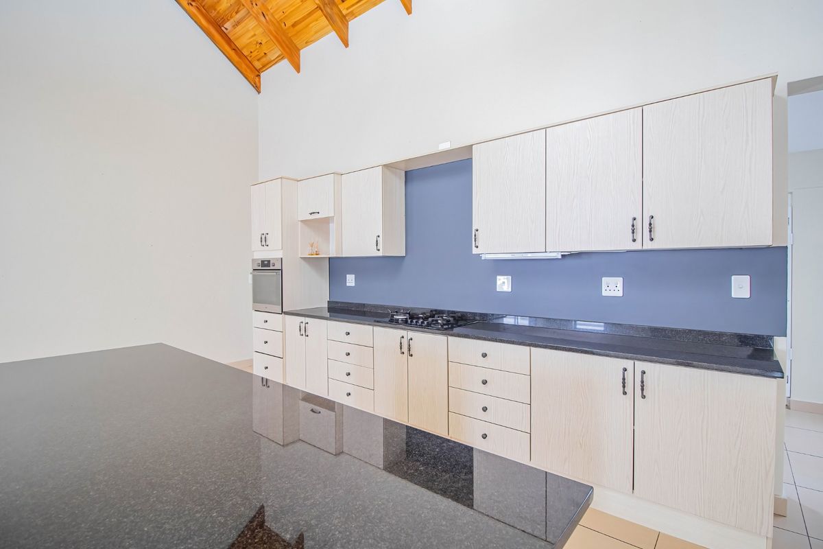 Modern kitchen featuring light wood cabinetry, dark granite countertops, built-in appliances, and high vaulted ceilings with exposed timber rafters.