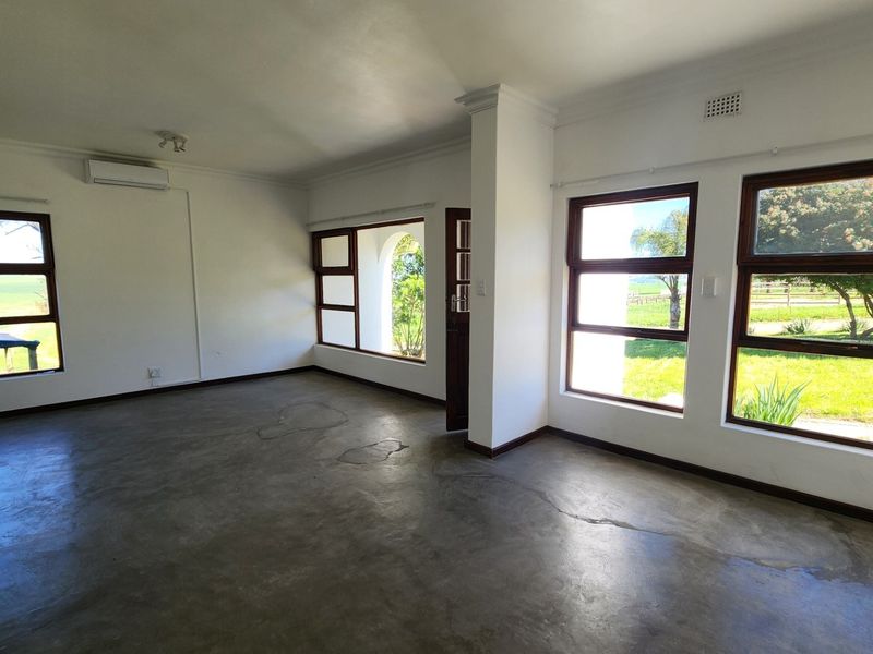 An empty living room featuring polished concrete floors, large timber-framed windows with garden views, and a wall-mounted air conditioning unit.