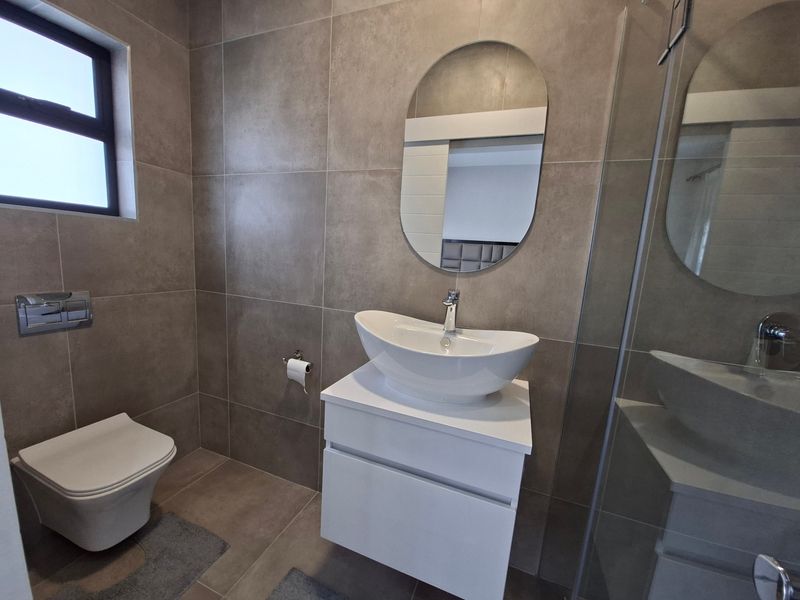 Modern bathroom featuring a wall-hung toilet, white vanity with vessel sink, oval mirror, and a glass-enclosed shower with neutral floor-to-ceiling tiling.