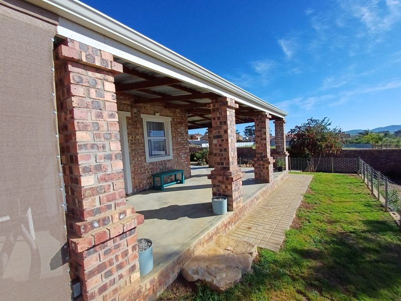 A covered outdoor patio featuring exposed brick columns, concrete flooring, and timber rafters, overlooking a landscaped garden area with paved pathways.