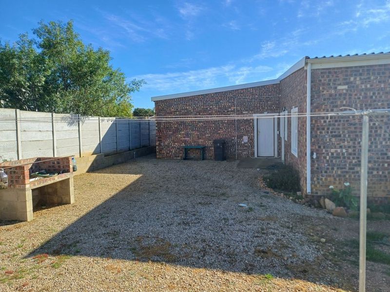 Rear exterior view of the property featuring a brick facade, gravel yard, and a built-in brick braai area enclosed by a concrete boundary wall.