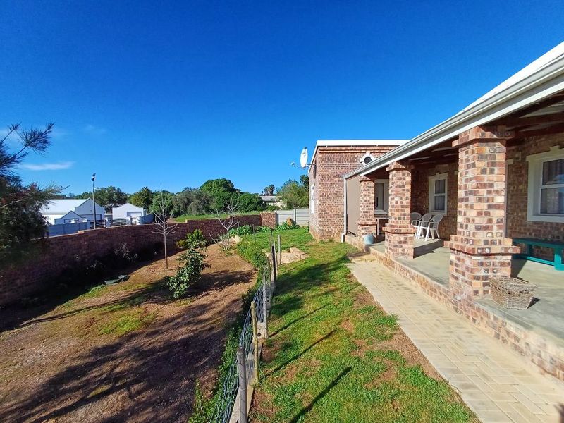 Rear view of the house featuring a covered brick patio, paved walkway, and a spacious garden enclosed by brick boundary walls and wire fencing.
