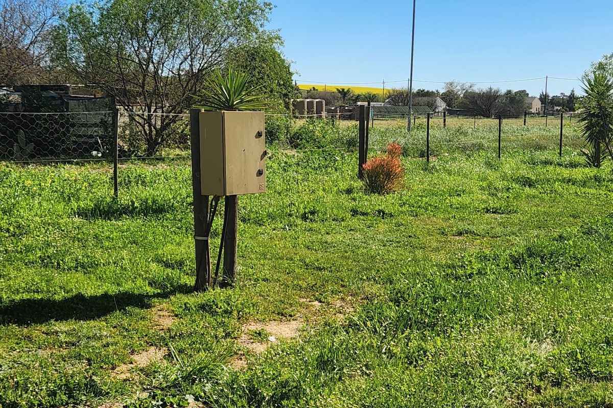 A view of a vacant residential plot featuring a grassy landscape, wire fencing, and an electrical distribution box.