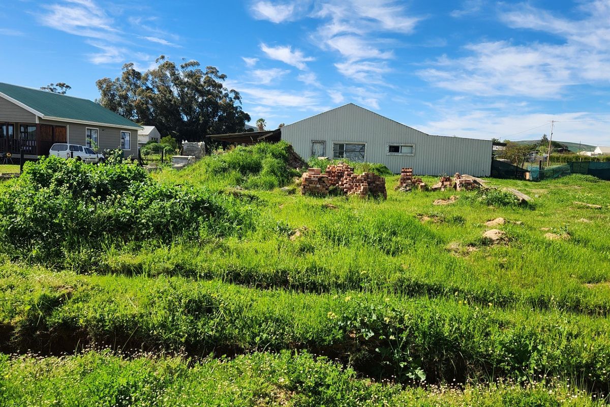 A view of a vacant residential plot featuring overgrown grass, scattered building materials, and an existing metal structure in the background.