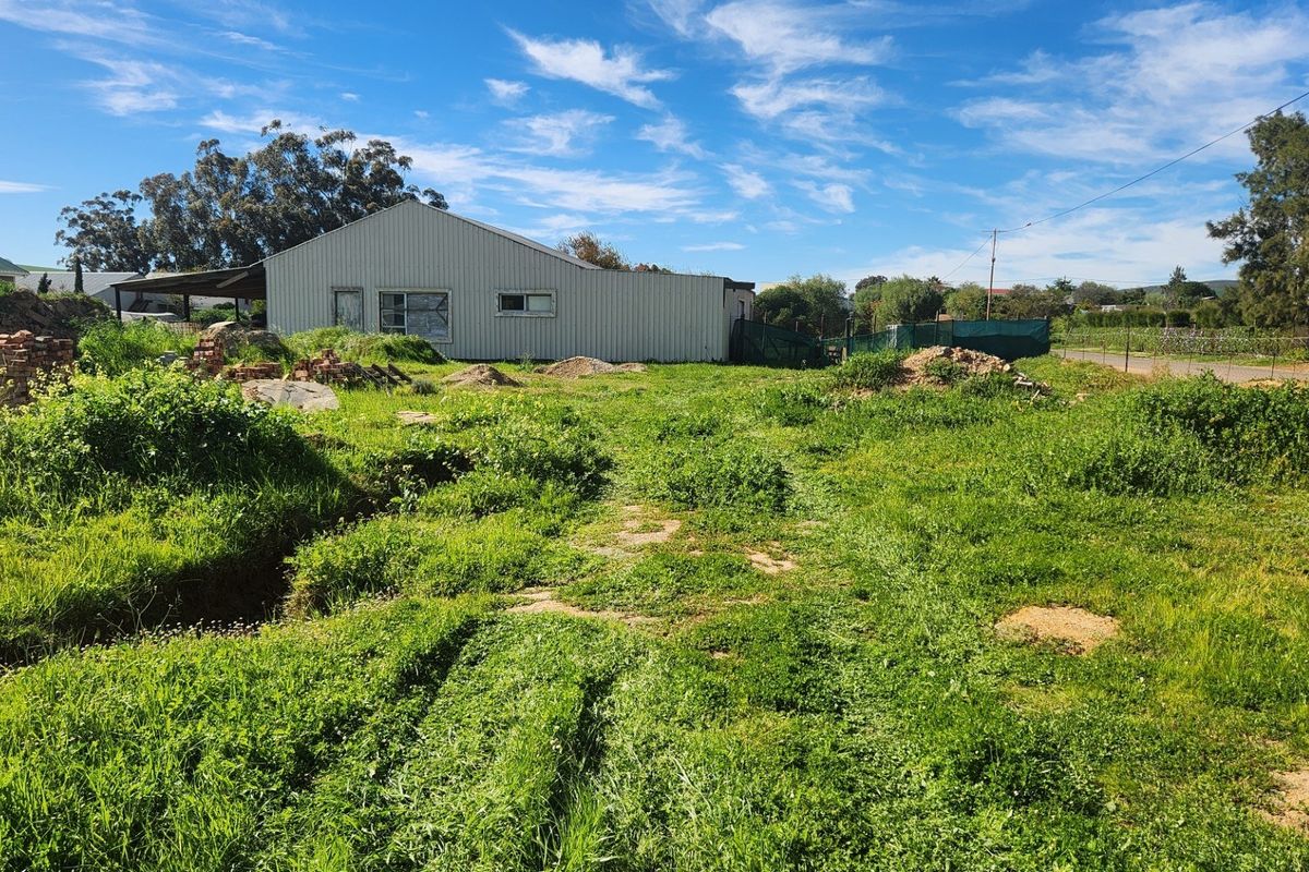 A view of a vacant residential plot featuring overgrown grass, piles of debris, and an existing metal shed structure on the property.