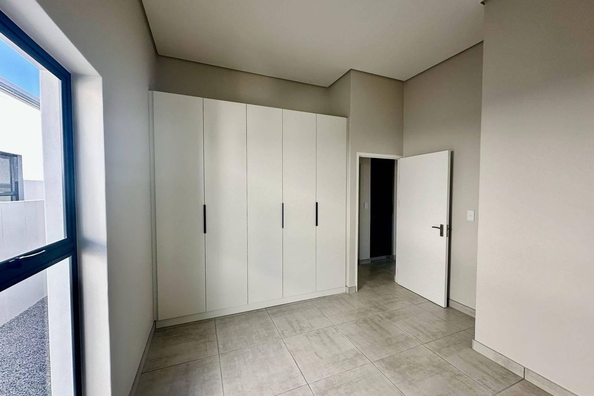 A bright, modern bedroom featuring neutral tiled flooring, built-in cupboards with sleek handles, and a large window providing natural light.