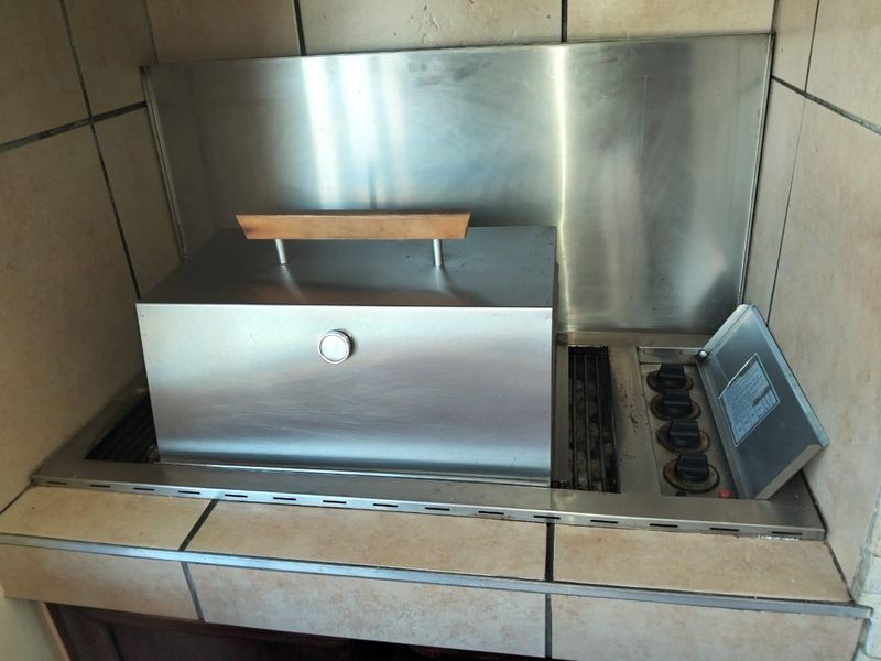 A built-in stainless steel braai unit set into a tiled countertop, featuring a metal splashback and integrated controls for outdoor cooking.