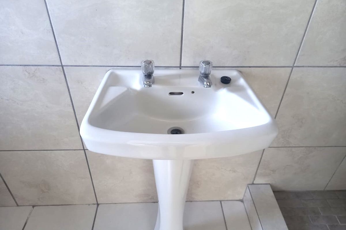 A clean, functional bathroom featuring a white pedestal basin with dual taps set against neutral ceramic wall tiles.