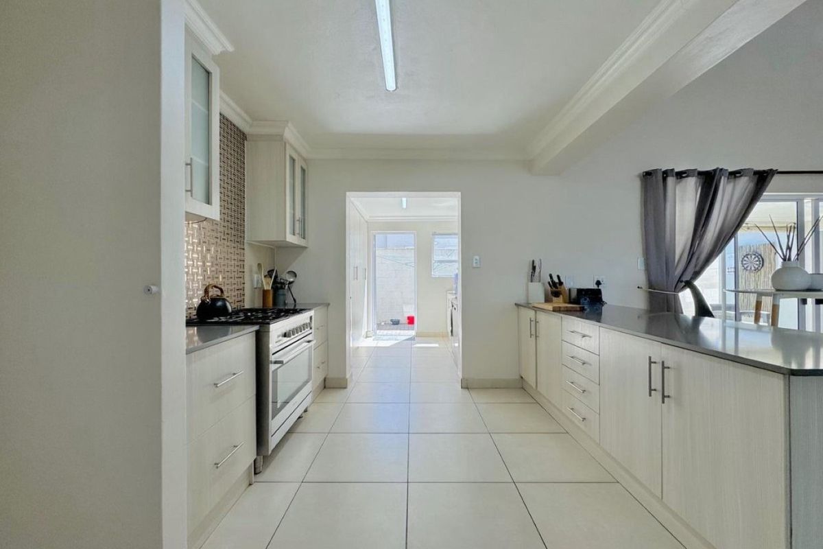 A clean, galley-style kitchen featuring light-toned cabinetry, stainless steel countertops, a gas stove, and a mosaic tile backsplash with tiled flooring throughout.
