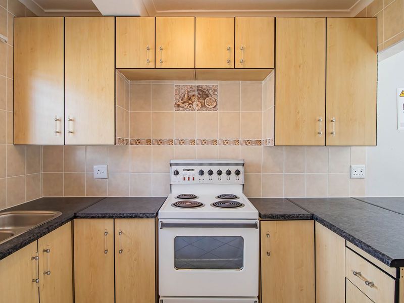 A functional kitchen featuring light wood-finish cabinetry, dark laminate countertops, a double stainless steel sink, and a freestanding electric stove with tiled backsplash.