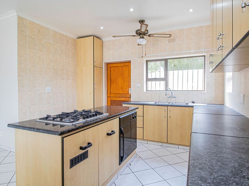 A functional kitchen featuring light wood cabinetry, dark granite countertops, a central island with a gas hob, and tiled flooring throughout.
