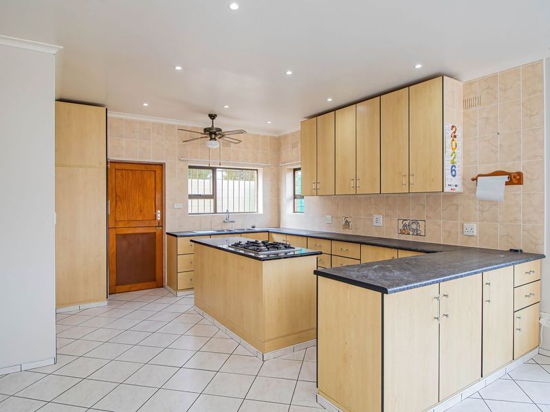A spacious kitchen featuring light wood cabinetry, dark laminate countertops, a central island with a gas hob, and tiled flooring throughout.