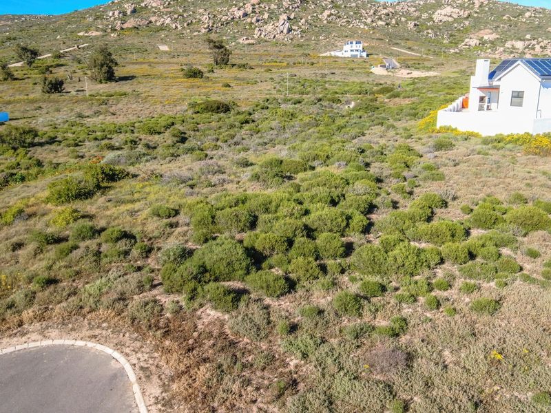 An aerial view of a vacant residential plot featuring natural vegetation, paved road access, and surrounding residential properties in a scenic landscape.
