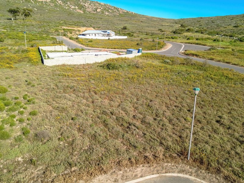 An elevated aerial view of a vacant residential plot featuring natural vegetation, adjacent paved roads, and established boundary walls in a scenic landscape.