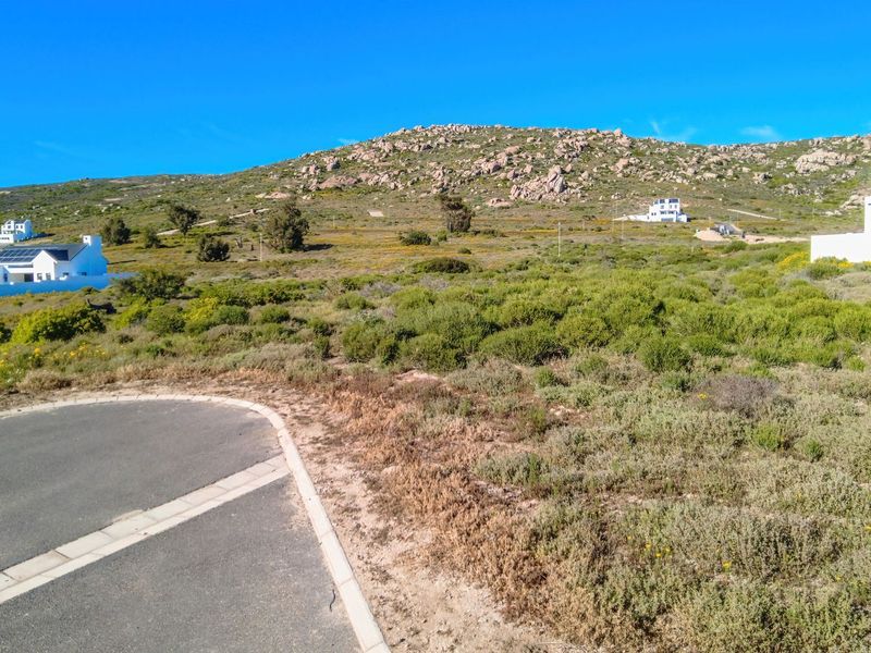 A vacant residential plot featuring natural coastal vegetation, adjacent to a paved road with kerbing, set against a scenic rocky hillside backdrop.