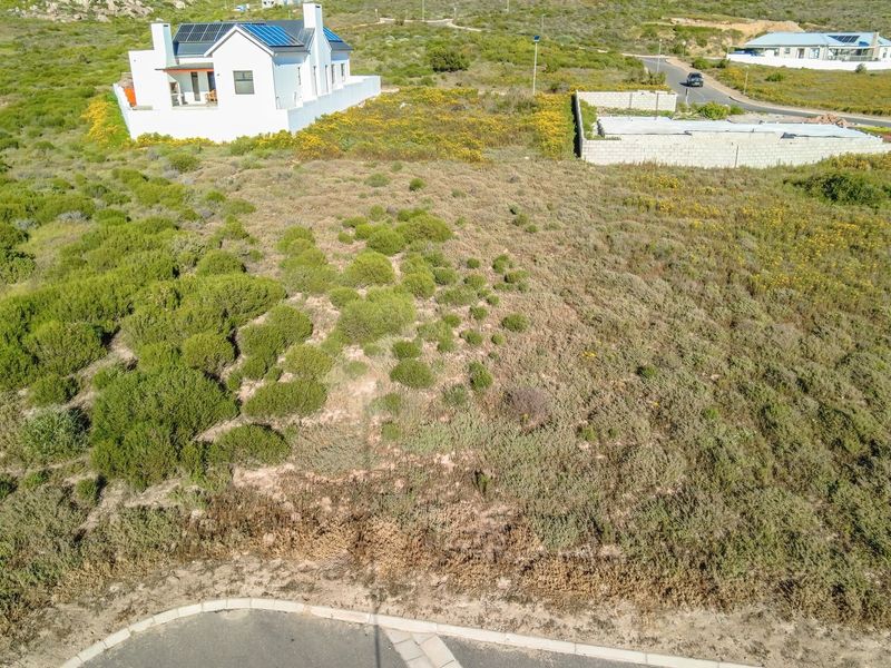 An aerial view of a vacant residential plot featuring natural vegetation, situated in a developed neighborhood with paved road access.