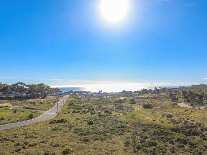 An aerial view of a vacant residential plot featuring natural coastal vegetation, paved road access, and distant ocean views.