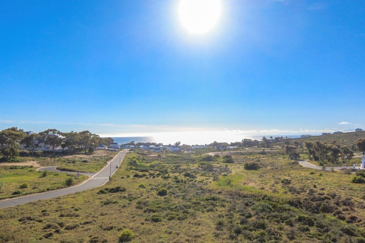 An aerial view of a vacant residential plot featuring natural coastal vegetation, paved road access, and distant ocean views.