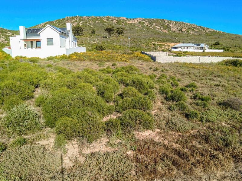 Aerial view of a residential plot featuring natural vegetation, a white house with solar panels, and a partially constructed concrete block boundary wall.