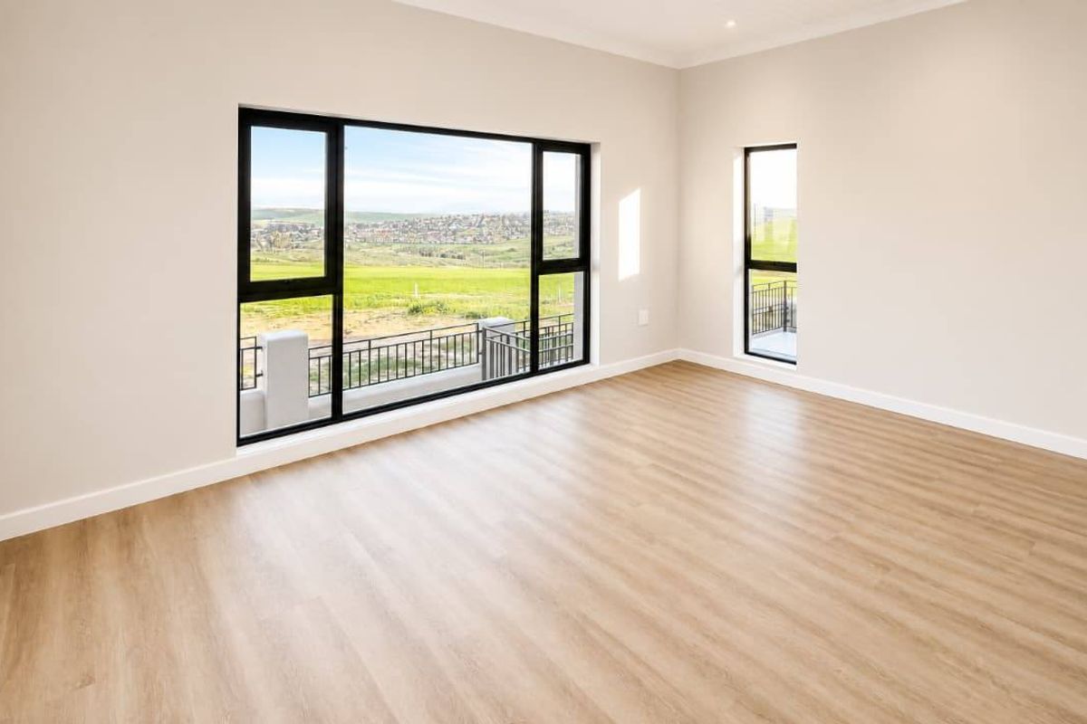 A bright, modern, and empty living area featuring light wood-look laminate flooring, neutral walls, and large windows offering scenic views.