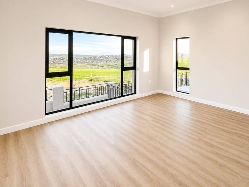 A bright, modern, and empty living area featuring light wood-look laminate flooring, neutral walls, and large windows offering scenic views.
