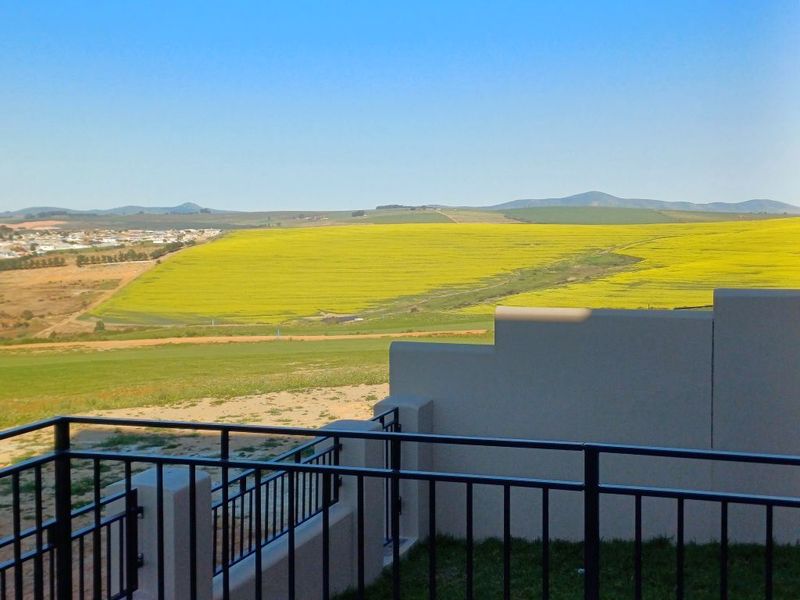 A scenic elevated view overlooking expansive yellow agricultural fields and distant hills, framed by a modern metal balustrade and masonry wall.