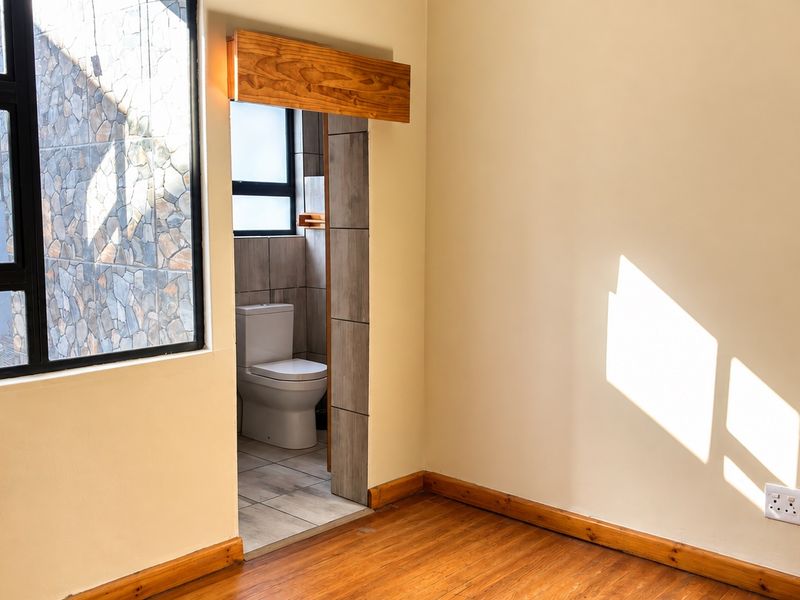 A bright, empty bedroom featuring warm wooden flooring, large aluminium-framed windows, and direct access to an en-suite bathroom.
