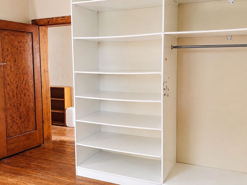 A bedroom interior featuring a built-in wardrobe with shelving and hanging space, complemented by wooden flooring and a classic wooden door.