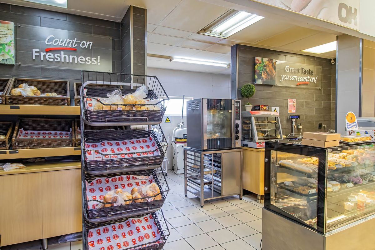 Commercial bakery or food service area featuring built-in display refrigeration, professional oven equipment, tiled flooring, and wall-mounted signage.