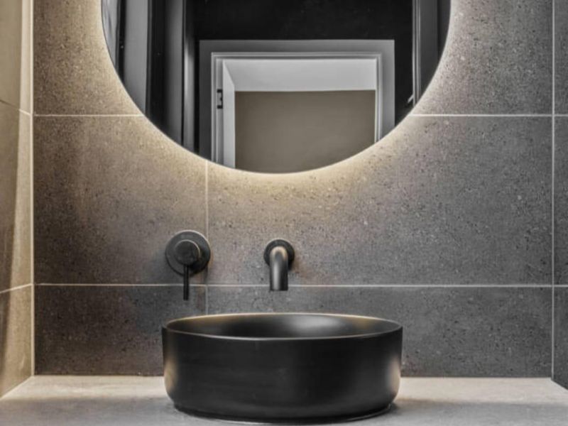 A modern, high-end bathroom vanity featuring a sleek vessel sink, wall-mounted matte black faucet, and a contemporary backlit circular mirror against dark stone tiling.