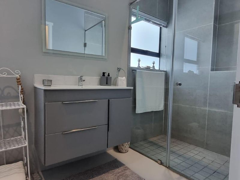 Modern bathroom featuring a grey vanity cabinet, basin, wall-mounted mirror, and a spacious glass-enclosed shower with tiled finishes.