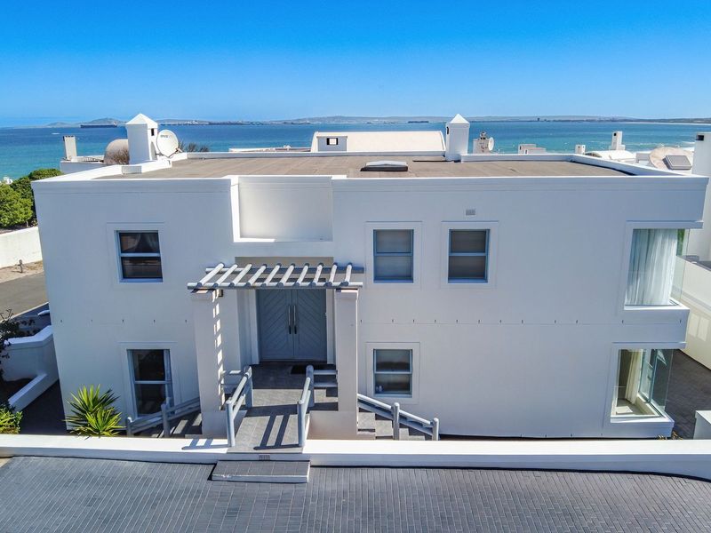 Aerial view of a modern white double-story house featuring a flat roof, pergola entrance, and paved driveway with ocean views.