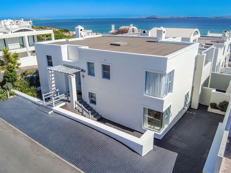 Aerial view of a modern two-story coastal residence featuring a flat roof, paved driveway, and a pergola entrance, situated near the ocean.