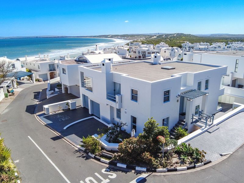 Aerial view of a modern white coastal house with a flat roof, paved driveway, and balconies, situated near the ocean.