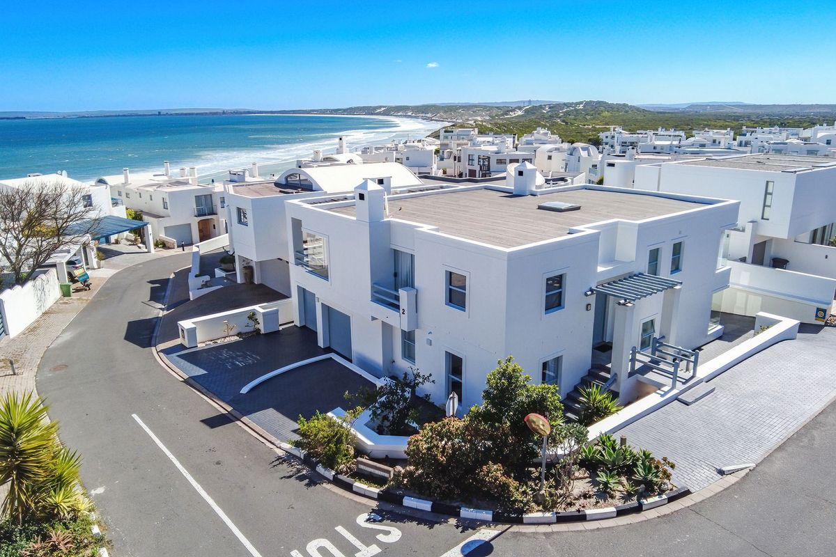 Aerial view of a modern white coastal house with a flat roof, paved driveway, and balconies, situated near the ocean.