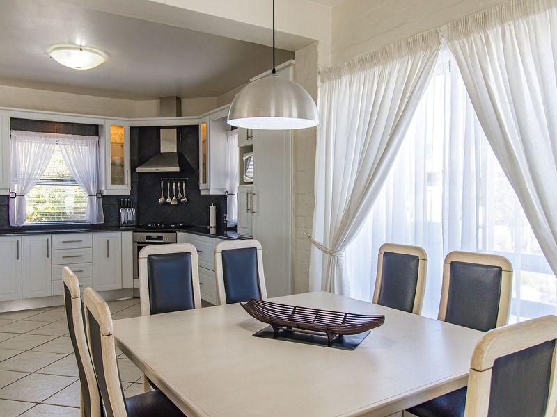 An open-plan kitchen and dining area featuring white cabinetry, dark countertops, tiled flooring, and natural light through large windows.