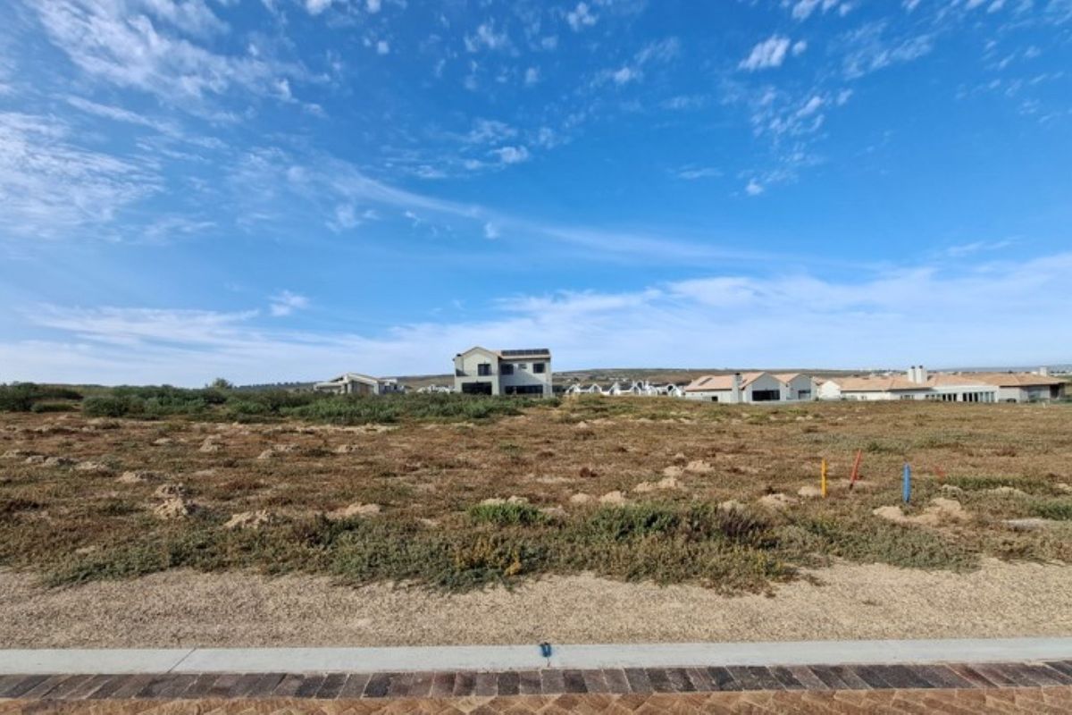 A vacant residential plot of land featuring natural vegetation, visible survey markers, and a paved curb, situated in a developed residential estate.