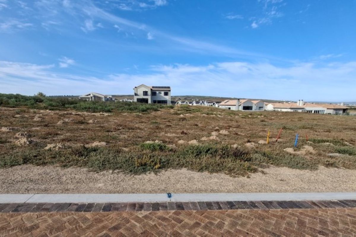 A vacant residential plot featuring natural vegetation and a paved verge, situated in a developed neighborhood.