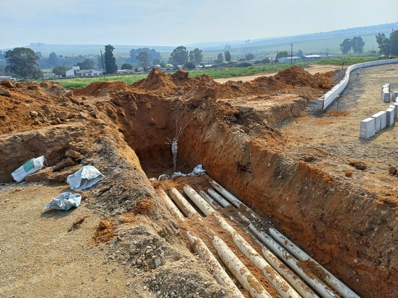 An active industrial construction site showing an excavated trench with installed underground piping and concrete curbing infrastructure.