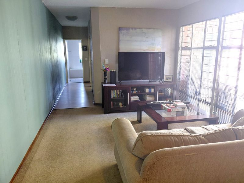 A living room area featuring carpet flooring, large windows with security bars, and direct access to a hallway leading to the bathroom.