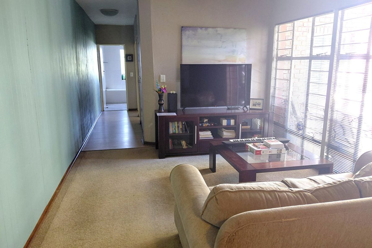 A living room area featuring carpet flooring, large windows with security bars, and direct access to a hallway leading to the bathroom.