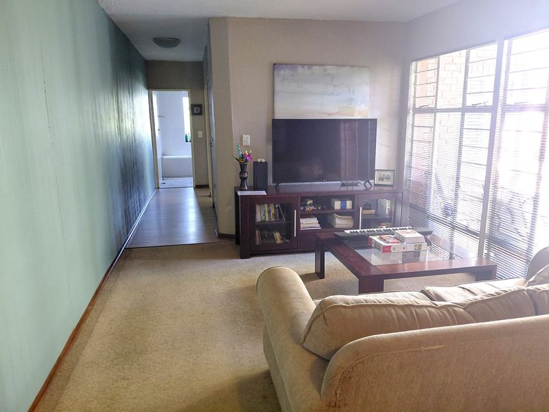 A living room area featuring carpet flooring and large windows with venetian blinds, providing access to the hallway and adjacent spaces.