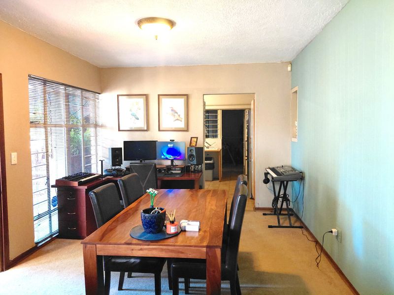 A multi-purpose living area featuring carpeted flooring, large windows with venetian blinds, and access to an adjacent utility space.
