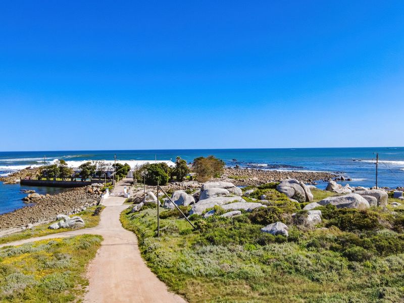 An aerial view of a coastal residential property featuring a dirt access road, natural rock formations, and direct proximity to the ocean.