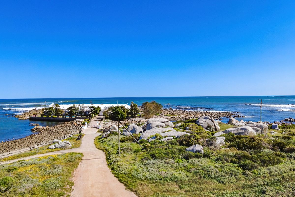 An aerial view of a coastal residential property featuring a dirt access road, natural rock formations, and direct proximity to the ocean.