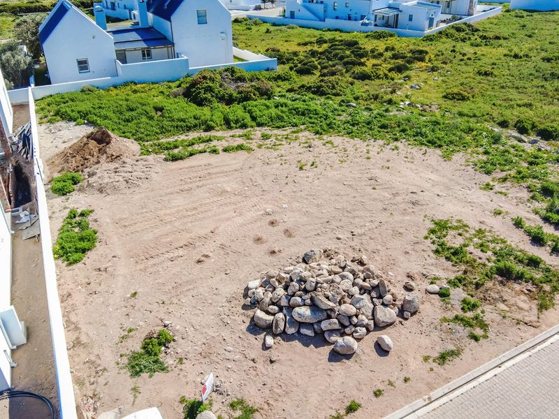 An aerial view of a vacant residential plot featuring cleared ground, piles of rocks, and soil, situated within a developed coastal residential estate.