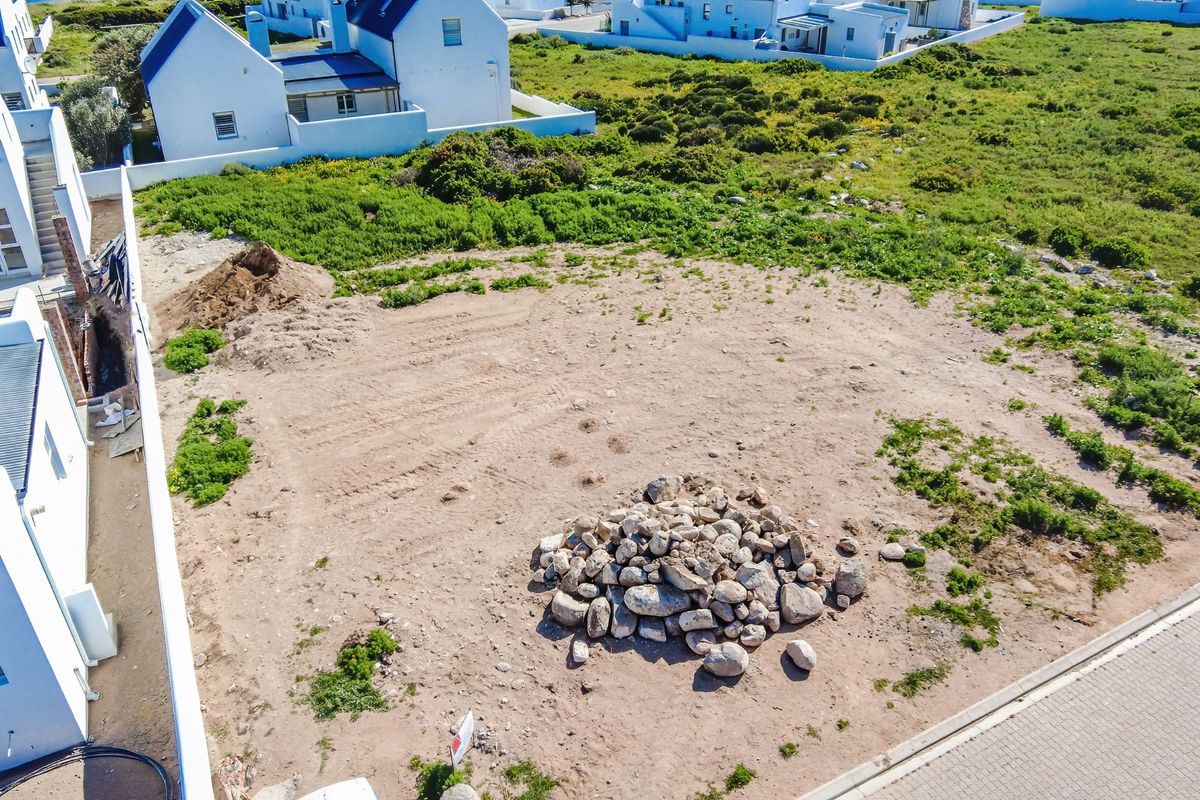 An aerial view of a vacant residential plot featuring cleared ground, piles of rocks, and soil, situated within a developed coastal residential estate.