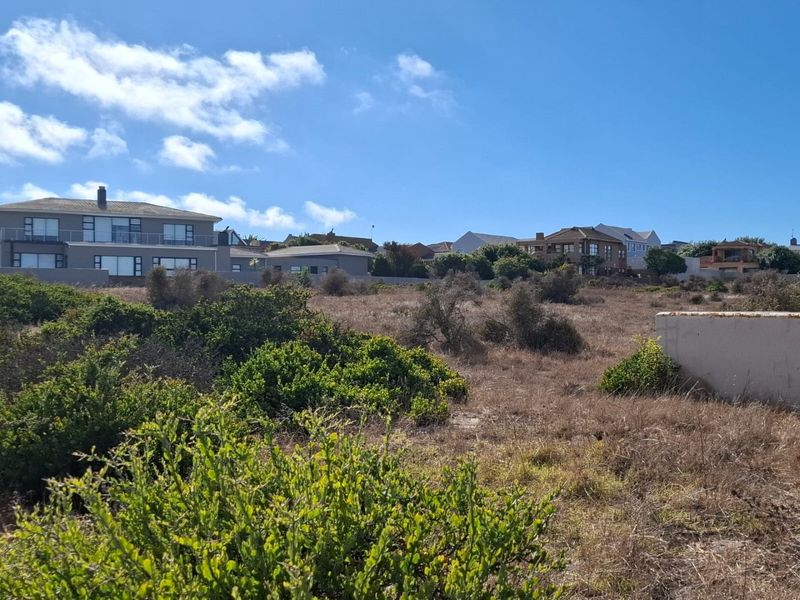 A view of a vacant residential plot featuring natural coastal vegetation and a partial retaining wall, with established homes visible in the background.