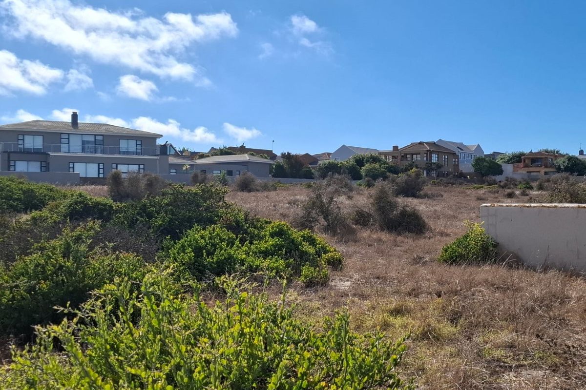 A view of a vacant residential plot featuring natural coastal vegetation and a partial retaining wall, with established homes visible in the background.