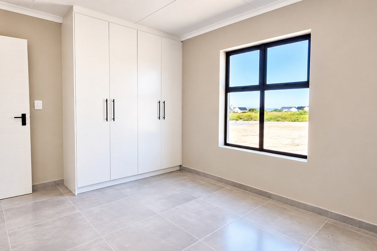 A bright, modern bedroom featuring neutral walls, large-format floor tiles, and ample built-in cupboard storage with a view through the window.